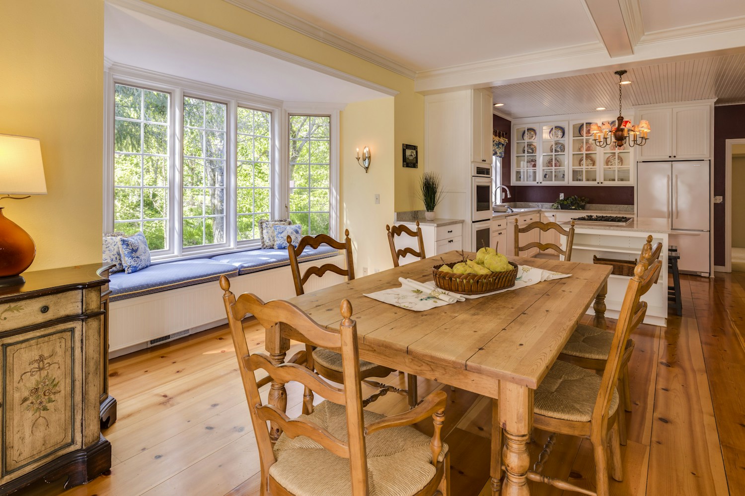 Sunlit dining room with a timber table, window seat and a bright open kitchen